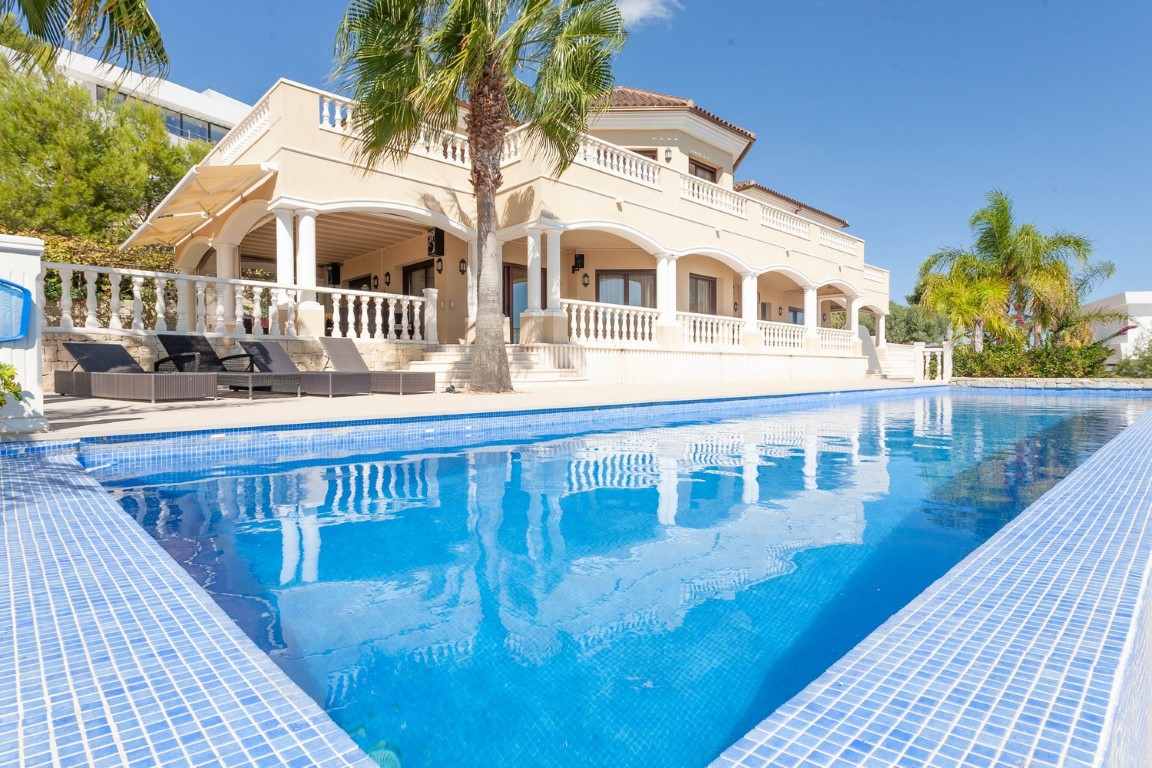 Large blue mosaic tile pool adjacent to a classic villa featuring arched covered terraces, white stone balustrades, and Mediterranean palm trees.