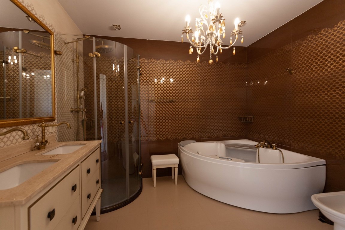 Master bathroom featuring a corner whirlpool tub, curved glass shower, and double vanity with brass fixtures. Textured brown wall tiles and a classic chandelier enhance the space.