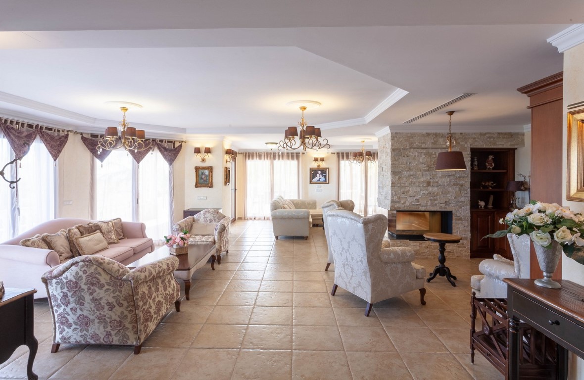 Spacious living room featuring ceramic tile flooring, a natural stone fireplace, and two distinct seating areas with classical furniture and chandeliers.