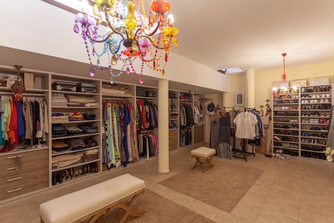 Spacious walk-in closet featuring open shelving, wood-finish drawers, and colorful crystal chandeliers. Includes upholstered benches, skylights, and a dedicated shoe display area.
