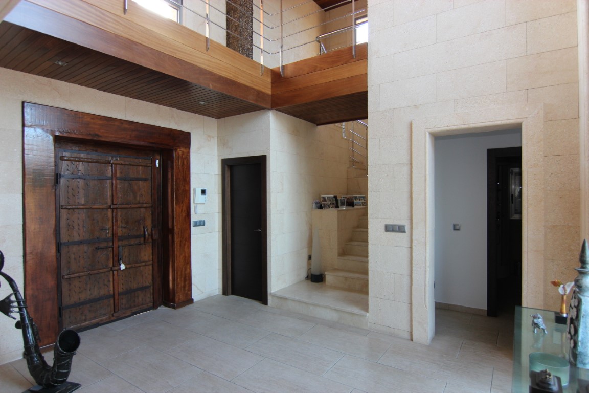 Double-height entrance hall featuring natural stone walls, a large rustic wooden door with iron details, and an upper gallery with wooden ceiling panels.