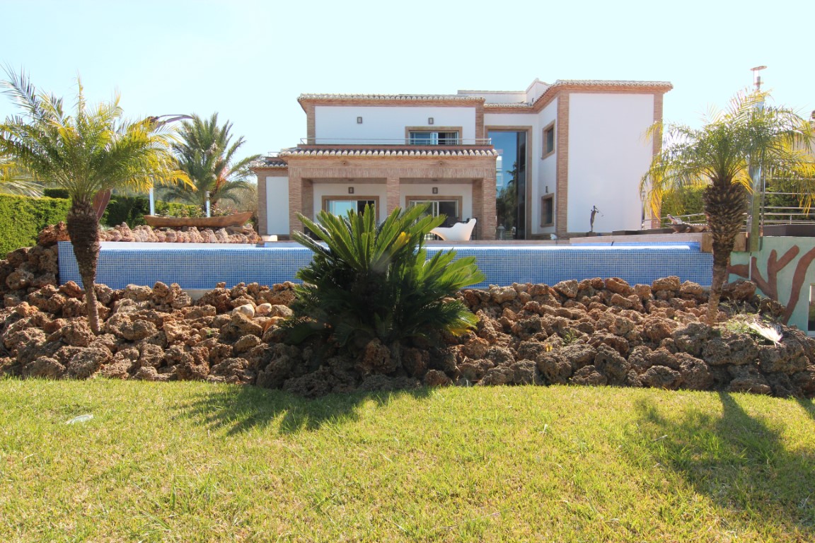 Exterior view featuring the blue mosaic pool and landscaped garden with volcanic rock, showcasing the villa's white facade and traditional brick accents.