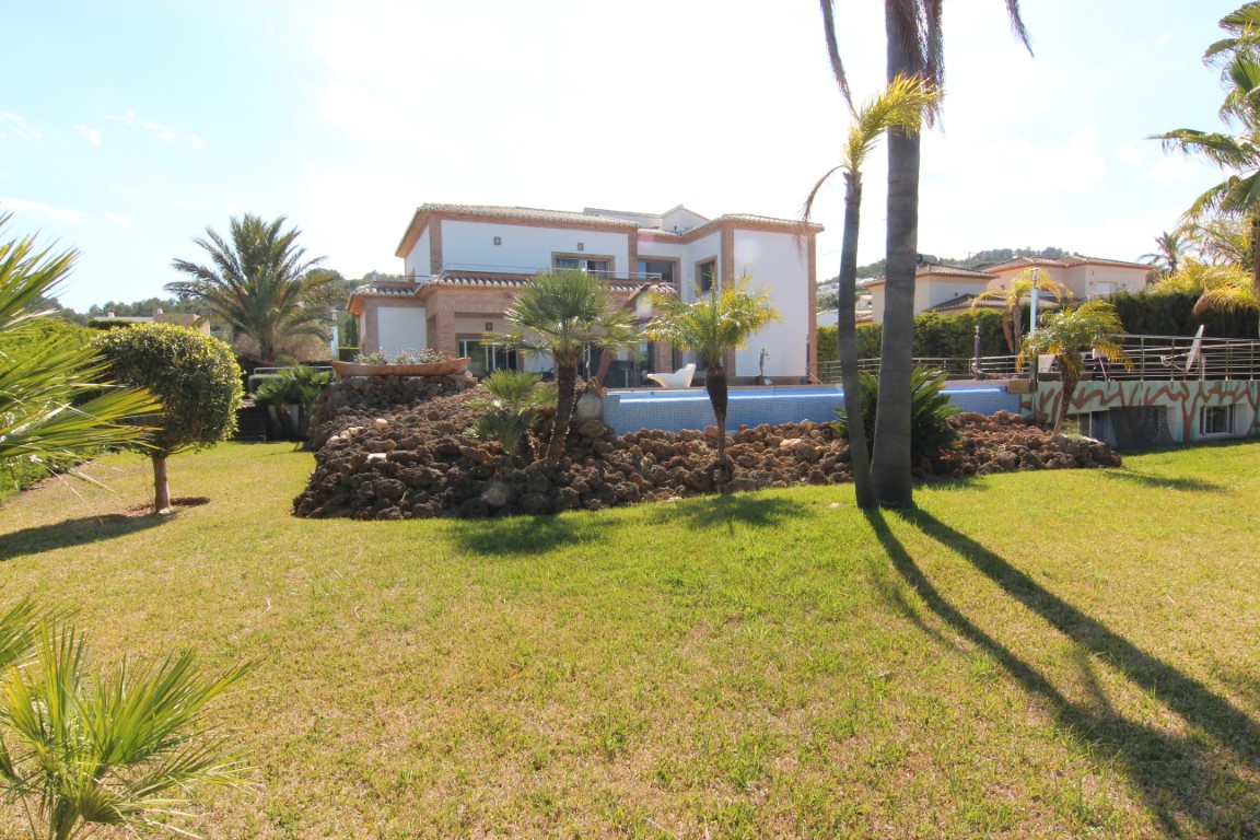 Exterior view of the villa featuring a manicured lawn, Mediterranean landscaping with palm trees, and an elevated swimming pool integrated into a natural stone rockery.