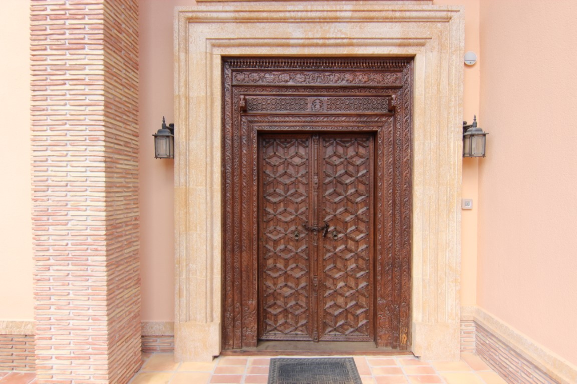 Hand-carved wooden entrance door featuring intricate geometric patterns, framed by natural stone and accented by traditional wall lanterns and exposed brickwork.
