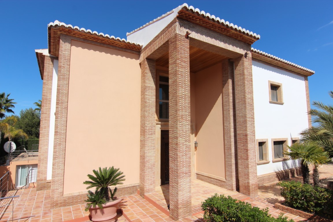 Main facade featuring a double-height entrance porch with slender brick columns, terracotta tiling, and a wooden ceiling under a clear blue sky.