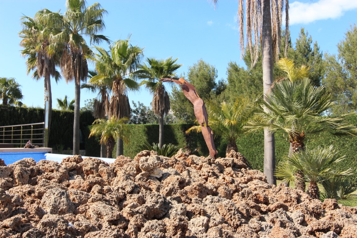 Mediterranean garden featuring various palm species, an abstract metal sculpture, and natural rock landscaping near the pool.