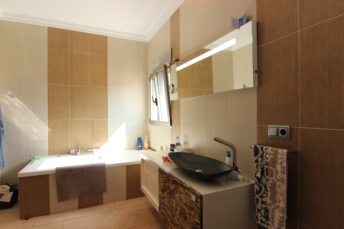 Modern bathroom featuring a built-in bathtub, a designer vessel sink, and wall tiling in alternating cream and wood-effect panels. Natural light enters through a side window.