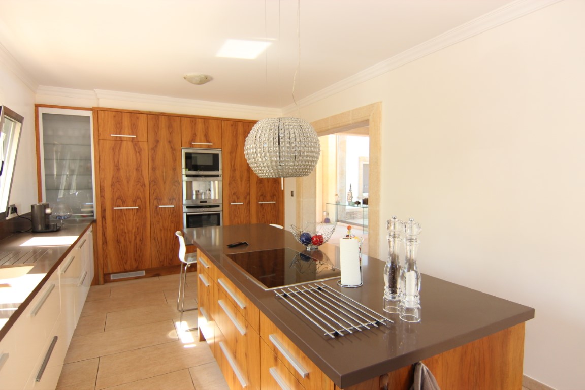 Modern kitchen with a central island, brown worktop, and natural wood cabinetry. Features integrated appliances, an induction hob, and a decorative crystal pendant light.