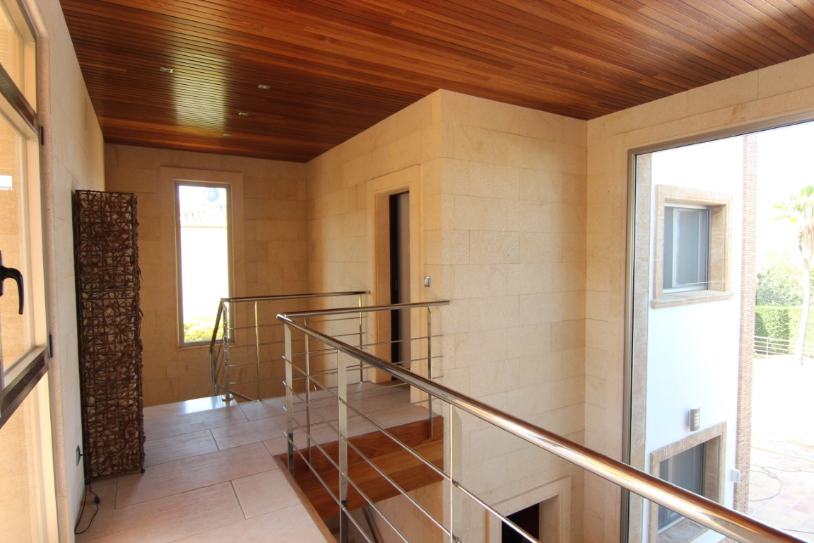 Upper floor landing featuring natural stone walls, wood-slatted ceilings, and stainless steel railings overlooking the double-height entrance.