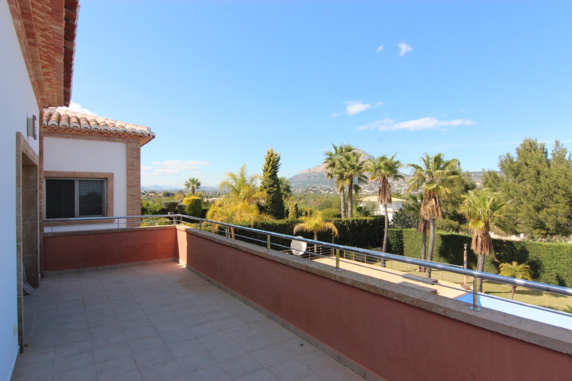 Upper-level terrace with stainless steel railings and stone trim, overlooking the landscaped garden, swimming pool, and Montgó mountain.