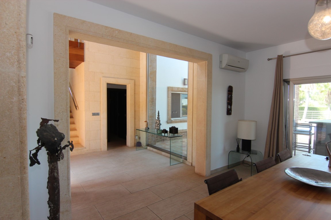 View from the dining area into the double-height entrance hall, featuring Tosca stone arches, a wooden staircase, and floor-to-ceiling windows.