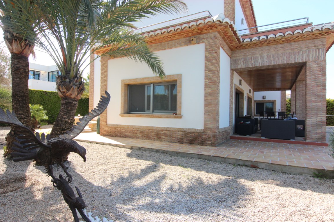 Villa exterior featuring brick accents, a covered porch with outdoor seating, and a gravel garden with palm trees and a metal eagle sculpture.