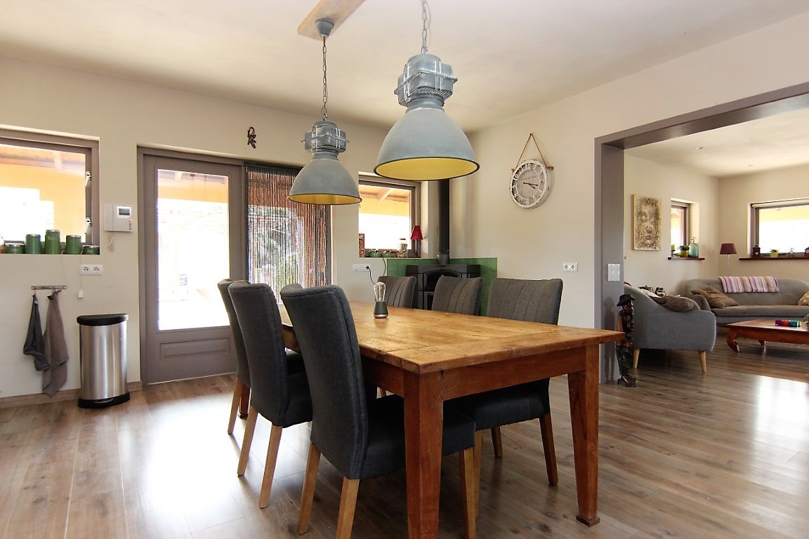 Dining area featuring a solid wood table, grey upholstered chairs, and industrial pendant lights, seamlessly connected to the living room and terrace.