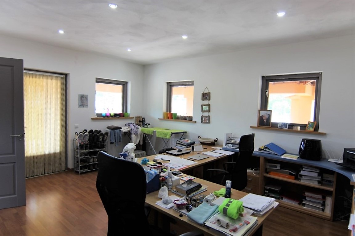 Functional home office featuring wood-effect flooring, recessed spotlights, and multiple windows. The space includes a large L-shaped desk and integrated shelving.