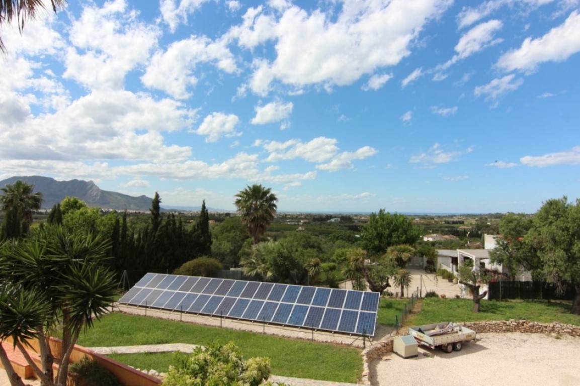 Panoramic view of the flat plot featuring a ground-mounted solar panel array, Mediterranean landscaping, and distant views of the mountains and coastline.