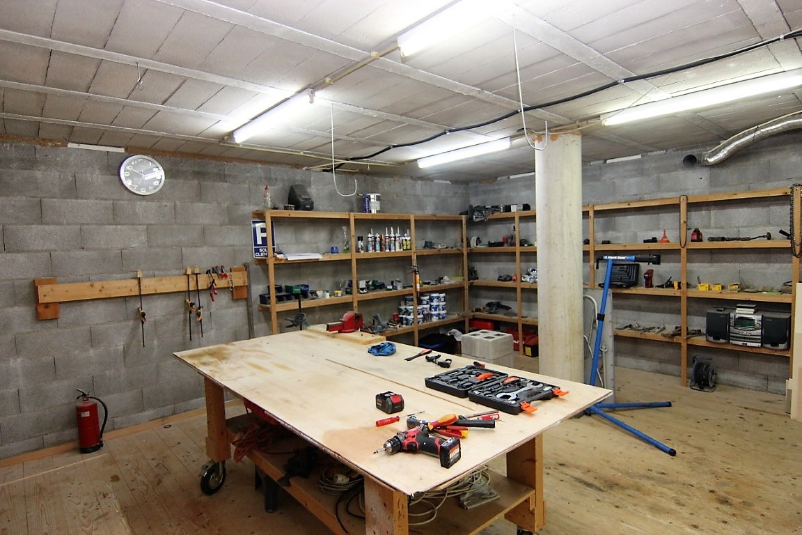 Spacious basement workshop featuring concrete block walls, perimeter wooden shelving, and a large central workbench with integrated tool storage.