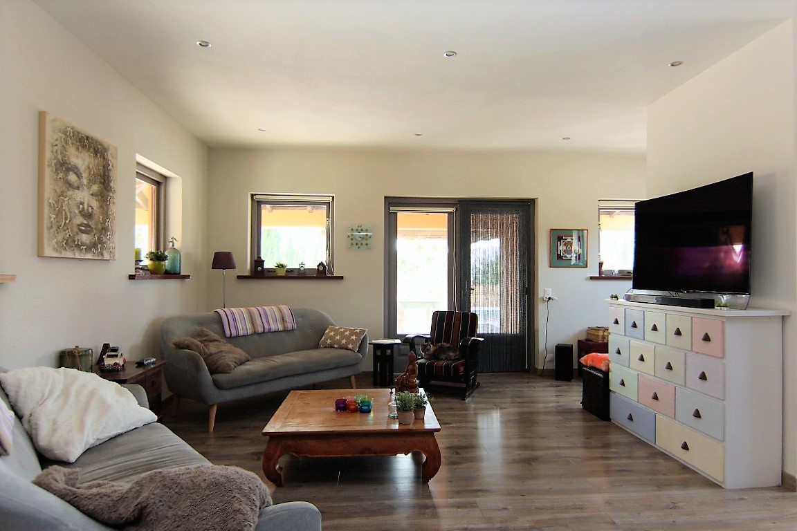 Spacious living room featuring wooden floors, grey sofas, and a colorful multi-drawer unit. Recessed ceiling lights and multiple windows provide ample natural light throughout the space.
