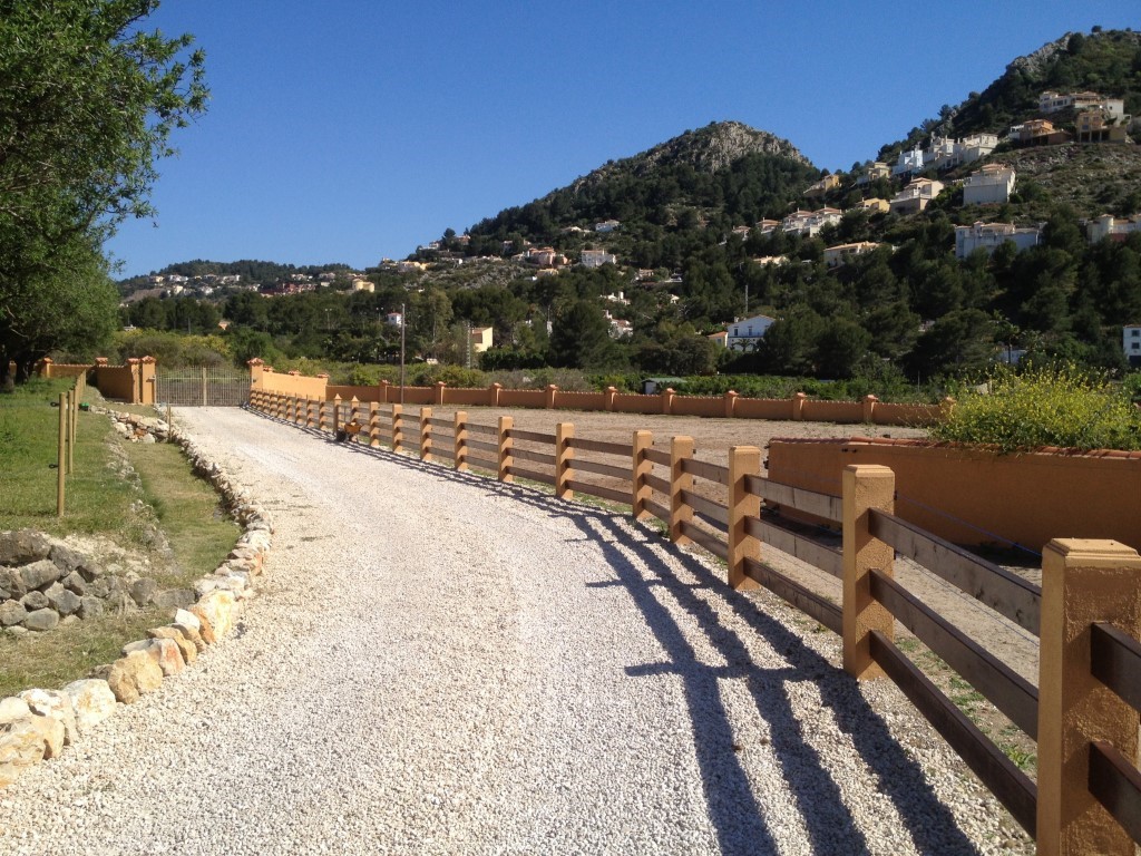 White gravel entrance path bordered by a wooden post-and-rail fence and perimeter walls, featuring clear views of the nearby hills and landscape.