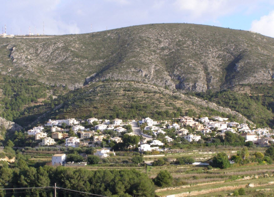 Panoramic view of a terraced hillside in Benitachell, featuring white residential villas nestled against a rugged mountain backdrop with native Mediterranean flora.