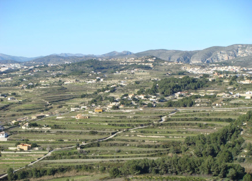 Panoramic view of terraced land in Benitachell, featuring stepped topography, Mediterranean vegetation, and distant mountain ranges under a clear sky.