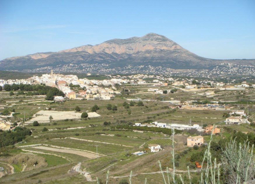 Panoramic view of the terraced plot in Benitachell, featuring the Montgó mountain massif and the traditional town center under a clear sky.