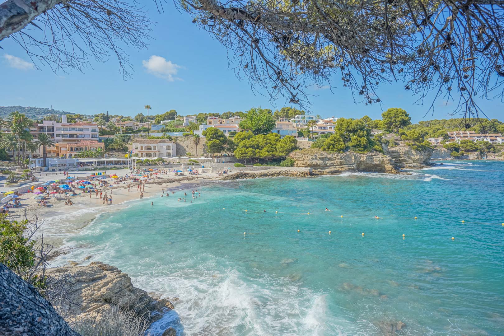 Panoramic view of Cala de la Fustera featuring turquoise waters, a sandy beach, and coastal residences framed by Mediterranean pine trees.
