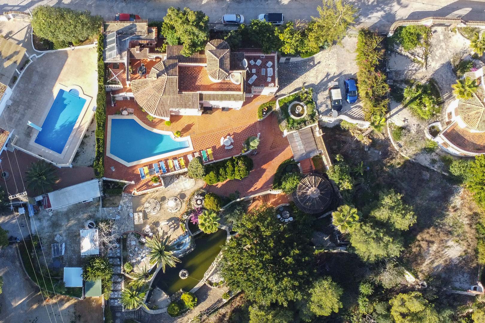 Aerial view of the property featuring the uniquely shaped swimming pool, expansive terracotta terraces, a large garden pond, and Mediterranean landscaping across the entire plot.