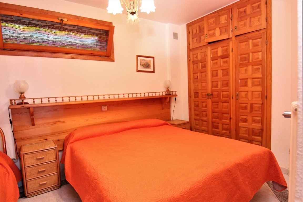 Bedroom featuring a wooden headboard with integrated shelving and built-in wardrobes. A unique horizontal stained-glass window provides character alongside wicker-detailed nightstands.