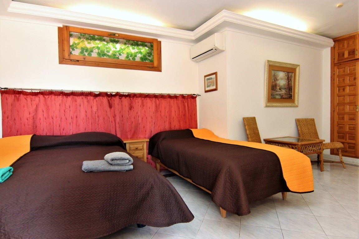 Bedroom featuring two beds with tiled flooring and air conditioning. The space includes wicker seating, a built-in wooden wardrobe, and indirect ceiling lighting.
