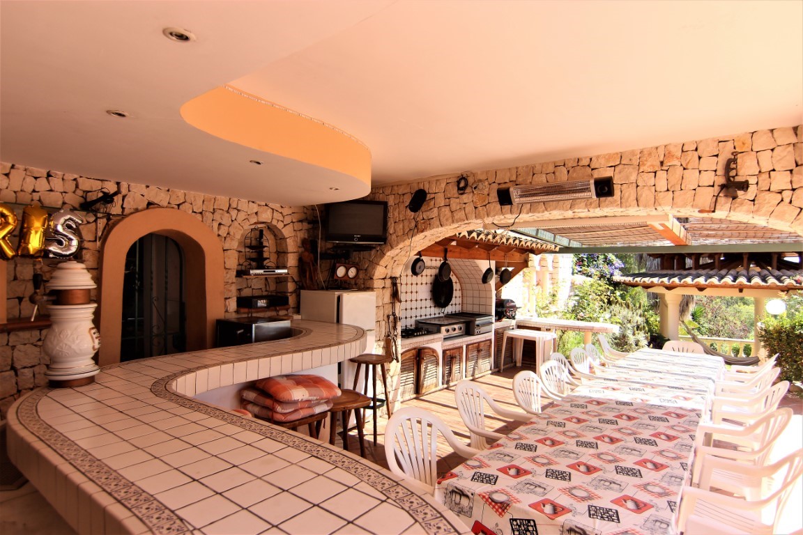Covered terrace featuring a tiled curved bar, professional outdoor kitchen, and large dining area set against rustic stone walls.