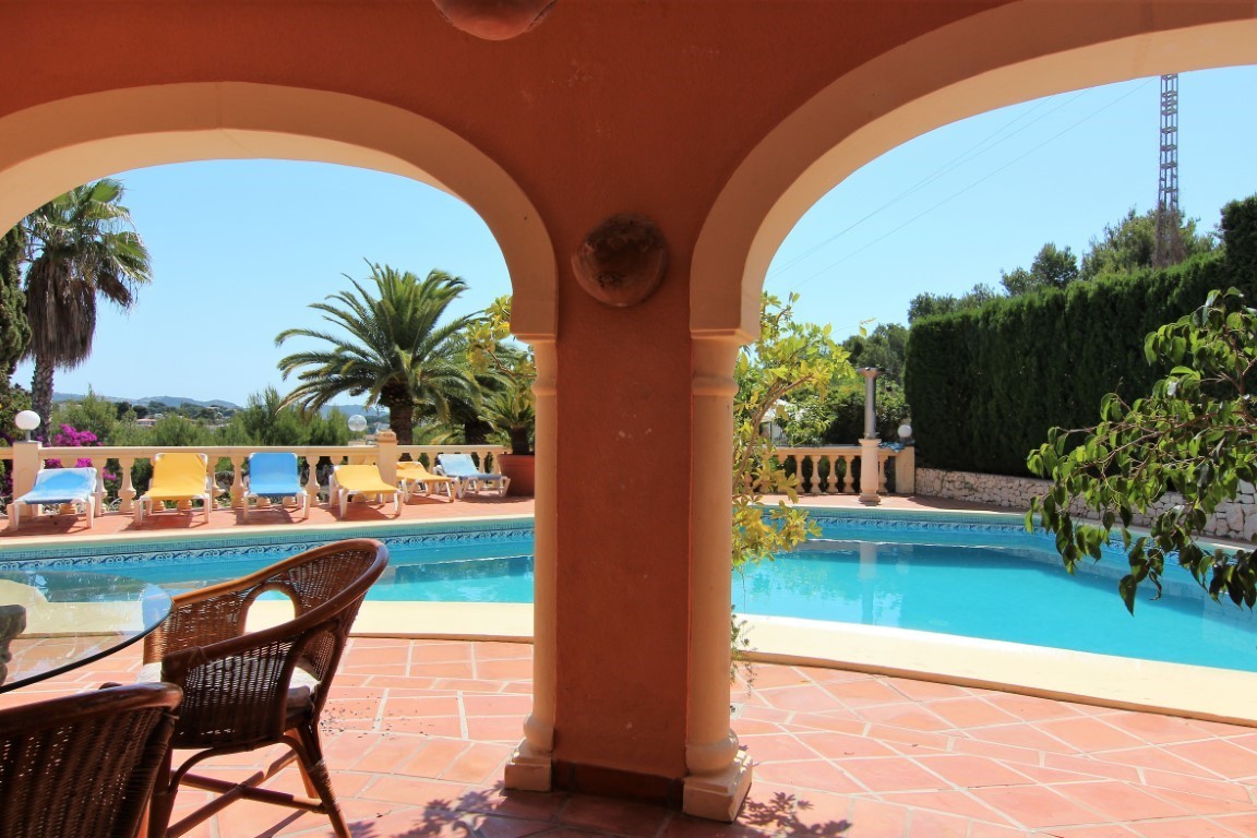 Covered terrace with Mediterranean arches overlooking a large swimming pool and sun deck with loungers, framed by palm trees and lush greenery.