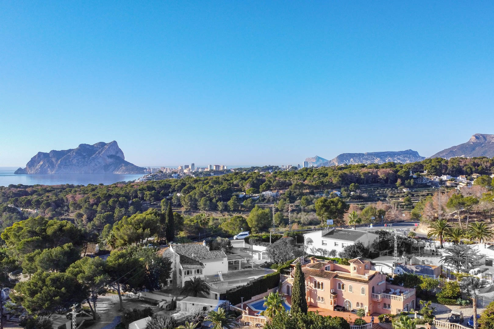 Elevated panoramic view of the Mediterranean coastline featuring the Peñón de Ifach, surrounded by lush pine forests and residential villas under a clear sky.