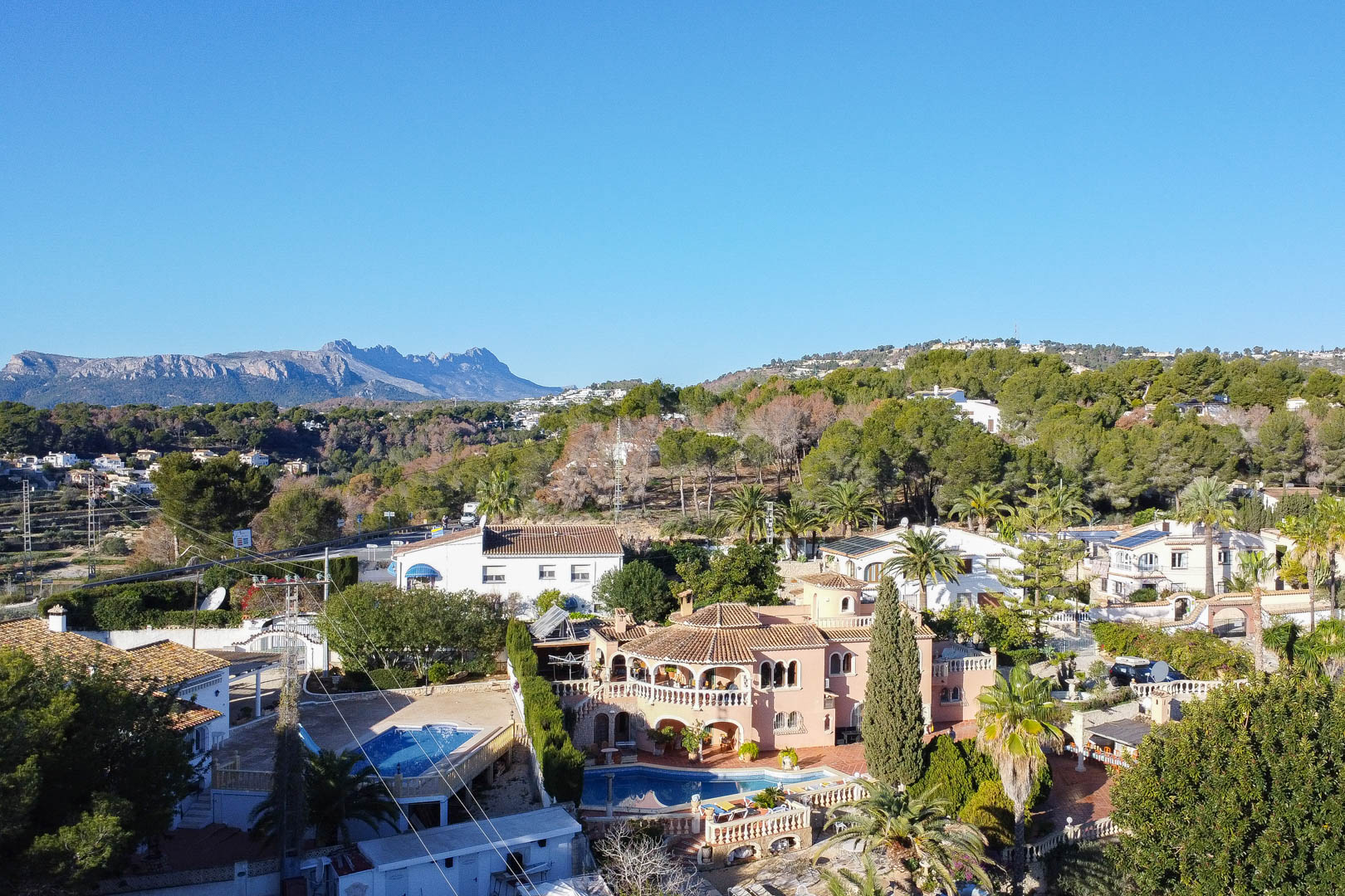 Elevated view of the Mediterranean villa featuring a curved swimming pool, arched terraces, and lush gardens set against a scenic mountain backdrop.