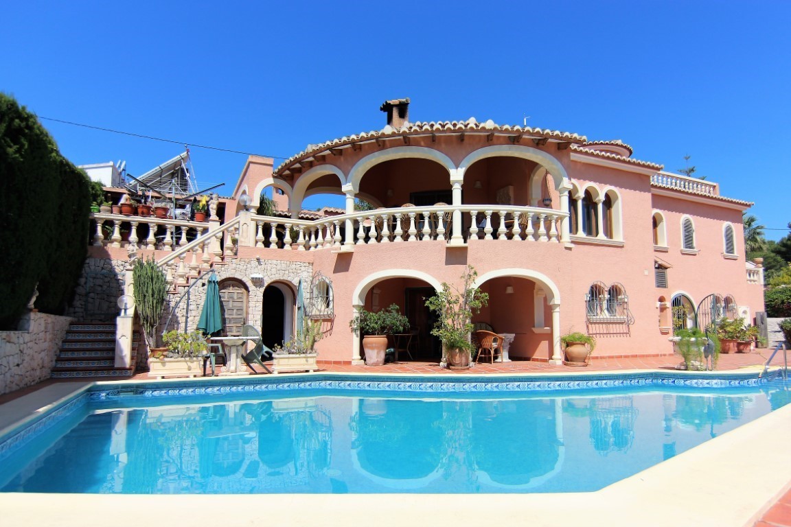 Exterior view of the villa featuring a large swimming pool, salmon-colored Mediterranean facade, arched terraces, and traditional stone balustrades.