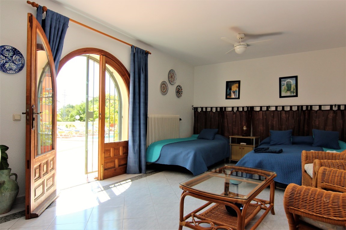 Ground floor bedroom featuring direct pool terrace access through an arched wooden door, furnished with wicker seating and a ceiling fan.