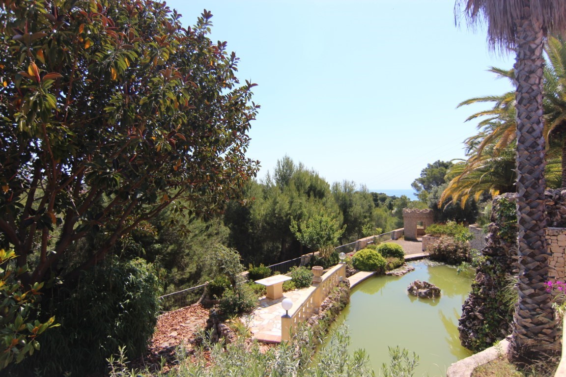 Large ornamental pond with a waterfall feature, surrounded by mature Mediterranean trees and stone walkways offering distant sea views.