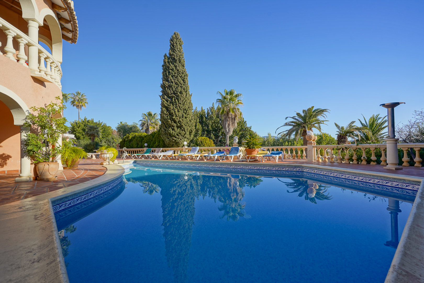 Large outdoor swimming pool featuring blue mosaic tiling, a terracotta terrace with stone balustrades, and a sunbathing area surrounded by Mediterranean greenery.