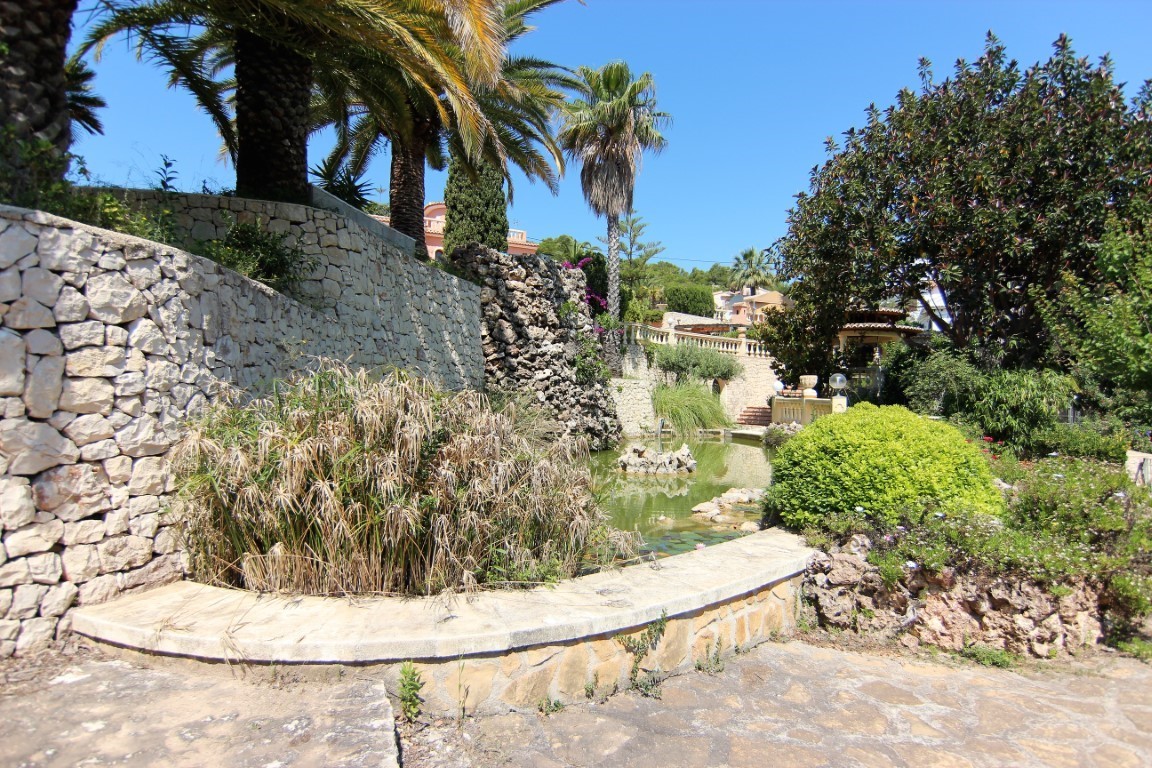 Mediterranean garden featuring an ornamental pond with a natural rock waterfall, dry stone walls, and mature palm trees under a clear sky.