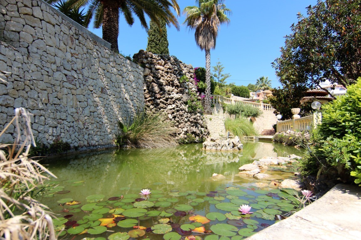 Ornamental garden pond featuring pink water lilies and a natural rock waterfall, set against dry stone walls and Mediterranean palms.