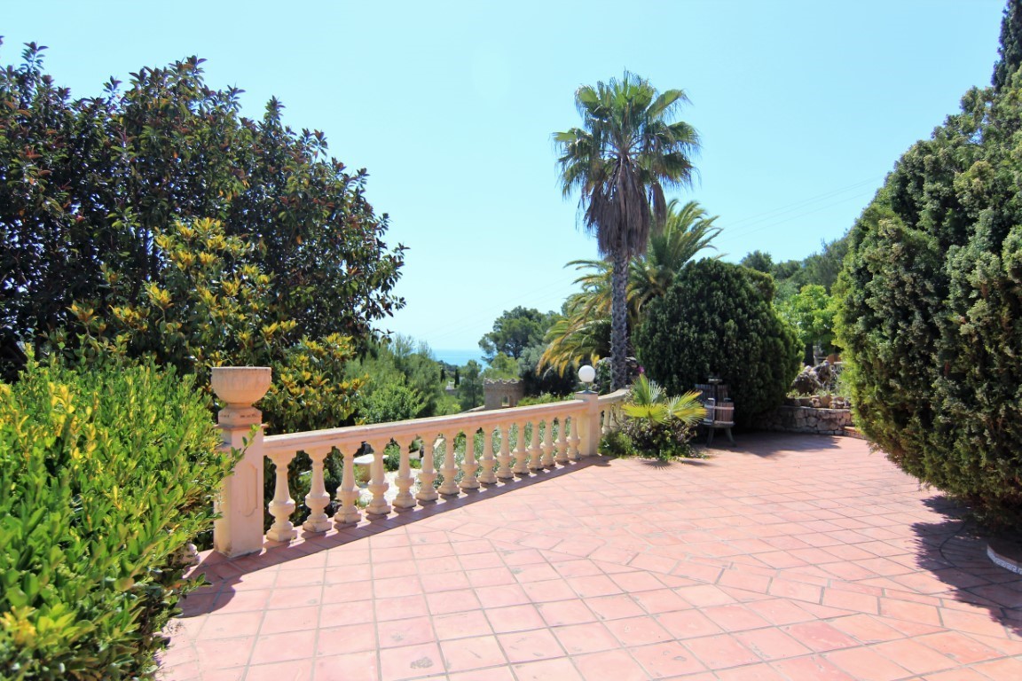 Outdoor terrace featuring terracotta tiling and a classic stone balustrade, bordered by mature Mediterranean greenery with distant sea views.