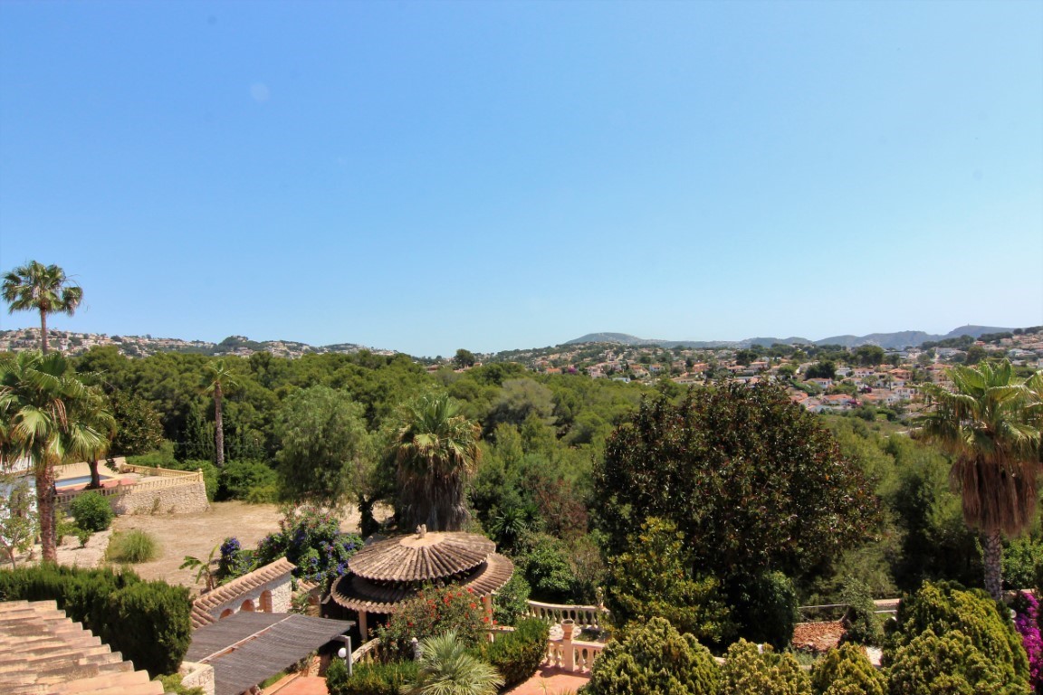 Panoramic view from the upper terrace overlooking a Mediterranean garden with palm trees, a tiered tiled gazebo, and distant residential hills under a clear sky.