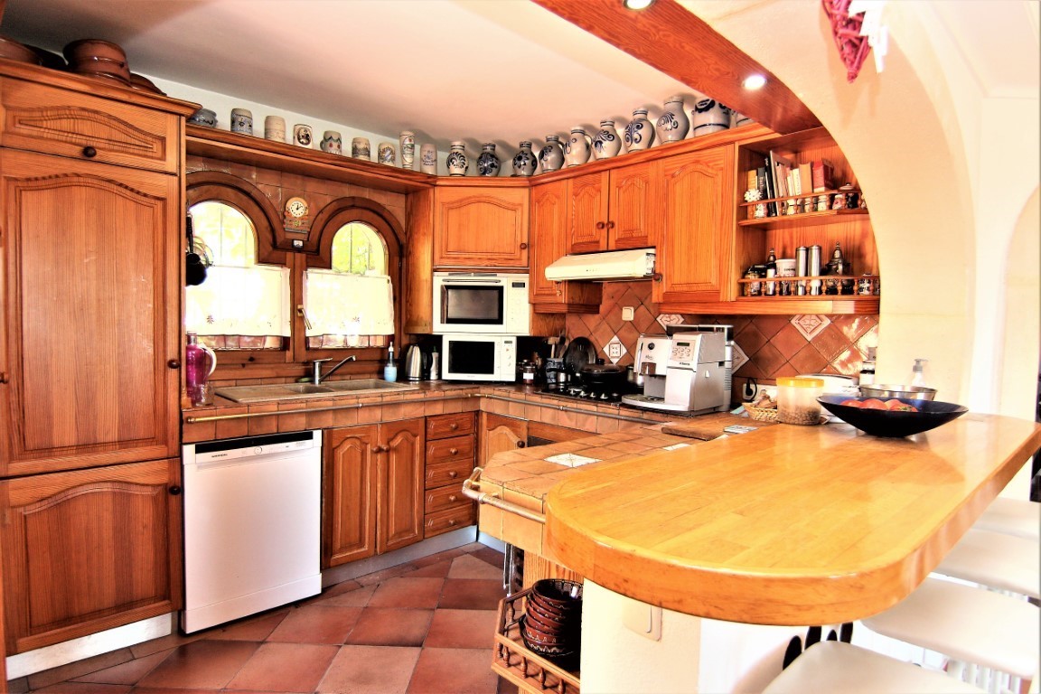 Rustic kitchen featuring wooden cabinetry, tiled countertops, and a breakfast bar. It includes integrated appliances, terracotta flooring, and traditional arched windows.