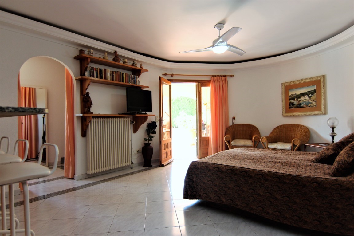 Spacious bedroom featuring direct terrace access, a wicker seating area, wooden shelving, and a ceiling fan over diagonal tiled flooring.