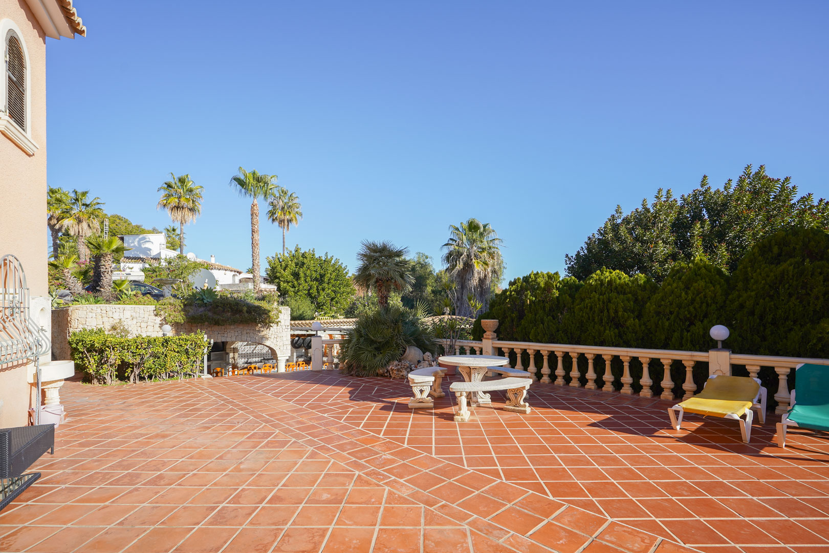 Spacious outdoor terrace featuring terracotta tiling, a stone balustrade, and a classic stone seating set, bordered by mature palms and lush Mediterranean greenery.