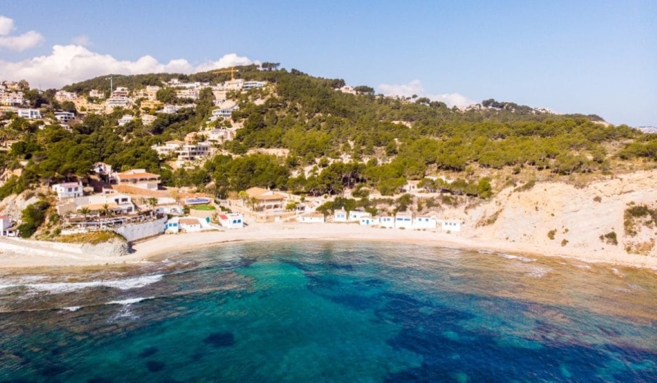 Aerial view of Cala de la Barraca featuring traditional white fishermen's houses, turquoise Mediterranean waters, and pine-covered hillsides.