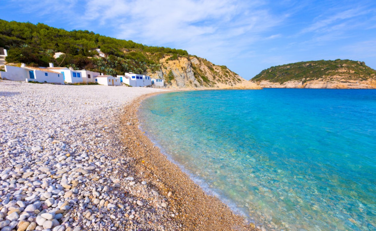 Panoramic view of Portichol beach in Jávea, featuring turquoise waters, white pebble shores, and traditional fisherman houses nestled against the rocky coastline.