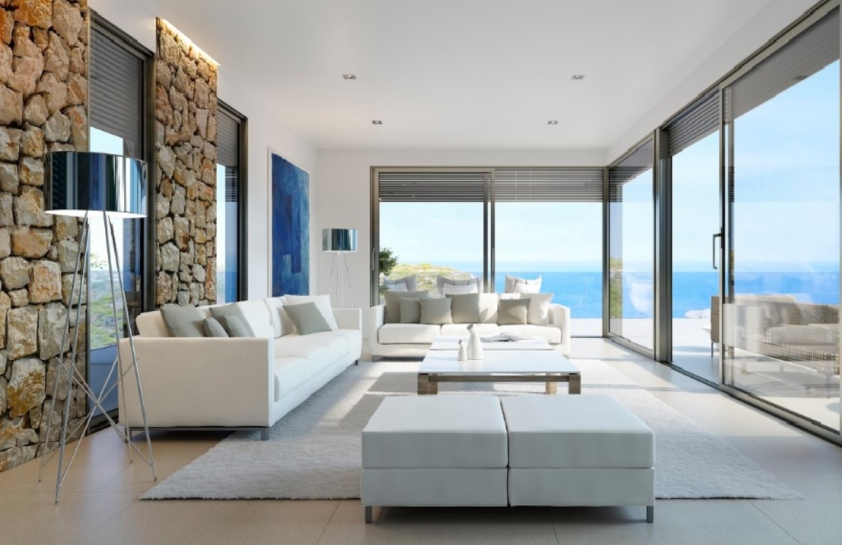 Bright living room featuring white sofas, a natural stone accent wall with LED lighting, and floor-to-ceiling glass doors overlooking the sea.