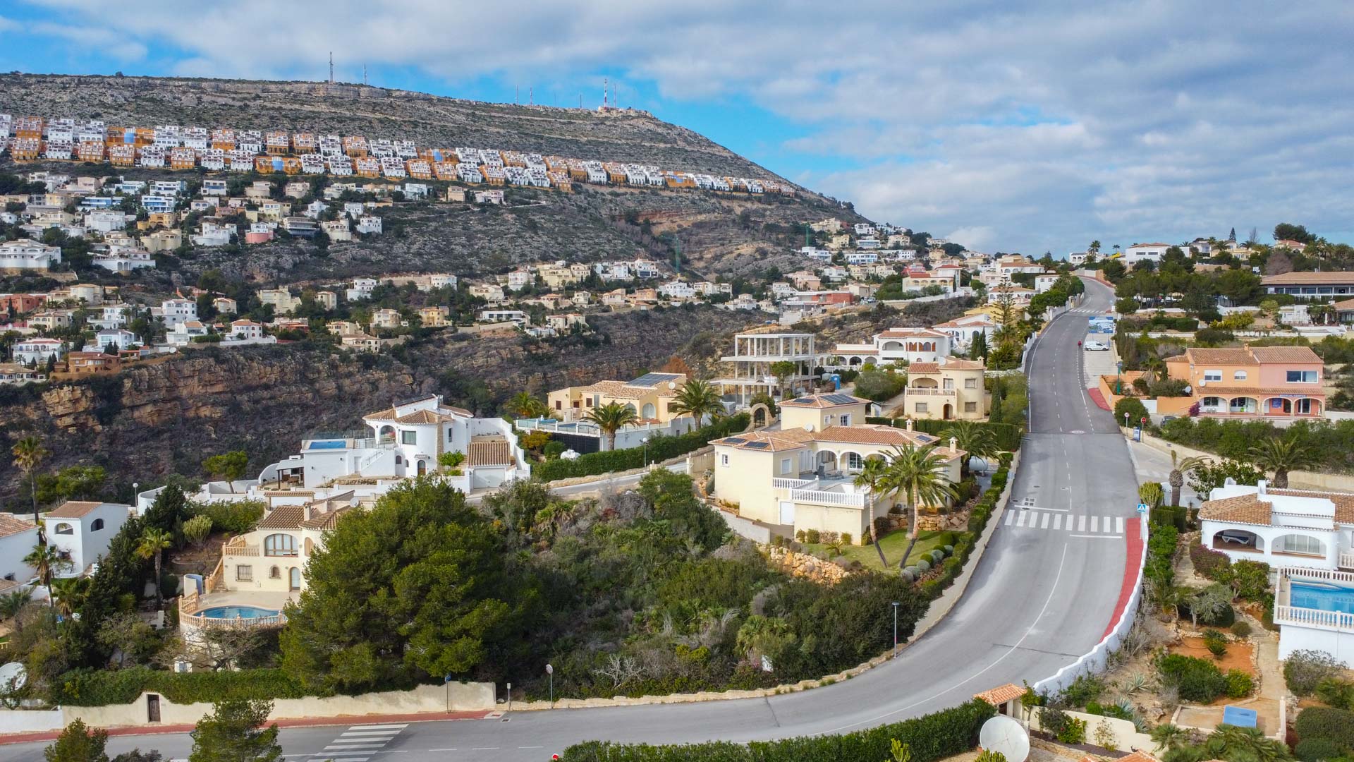 Aerial view of the Cumbre del Sol residential area, featuring winding roads, Mediterranean villas with terracotta roofs, and terraced housing along the hillside.