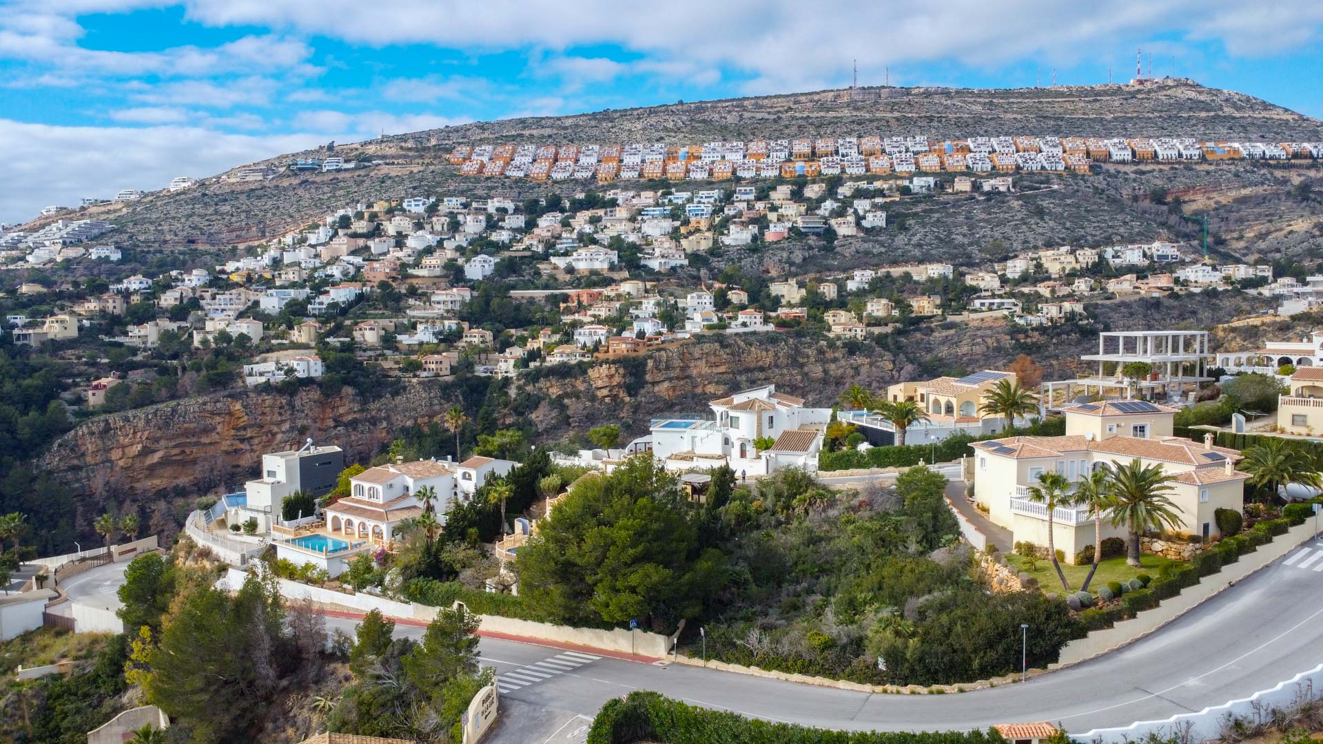 Elevated drone view of the Cumbre del Sol residential area, featuring hillside villas with Mediterranean architecture surrounded by green landscapes and winding roads.