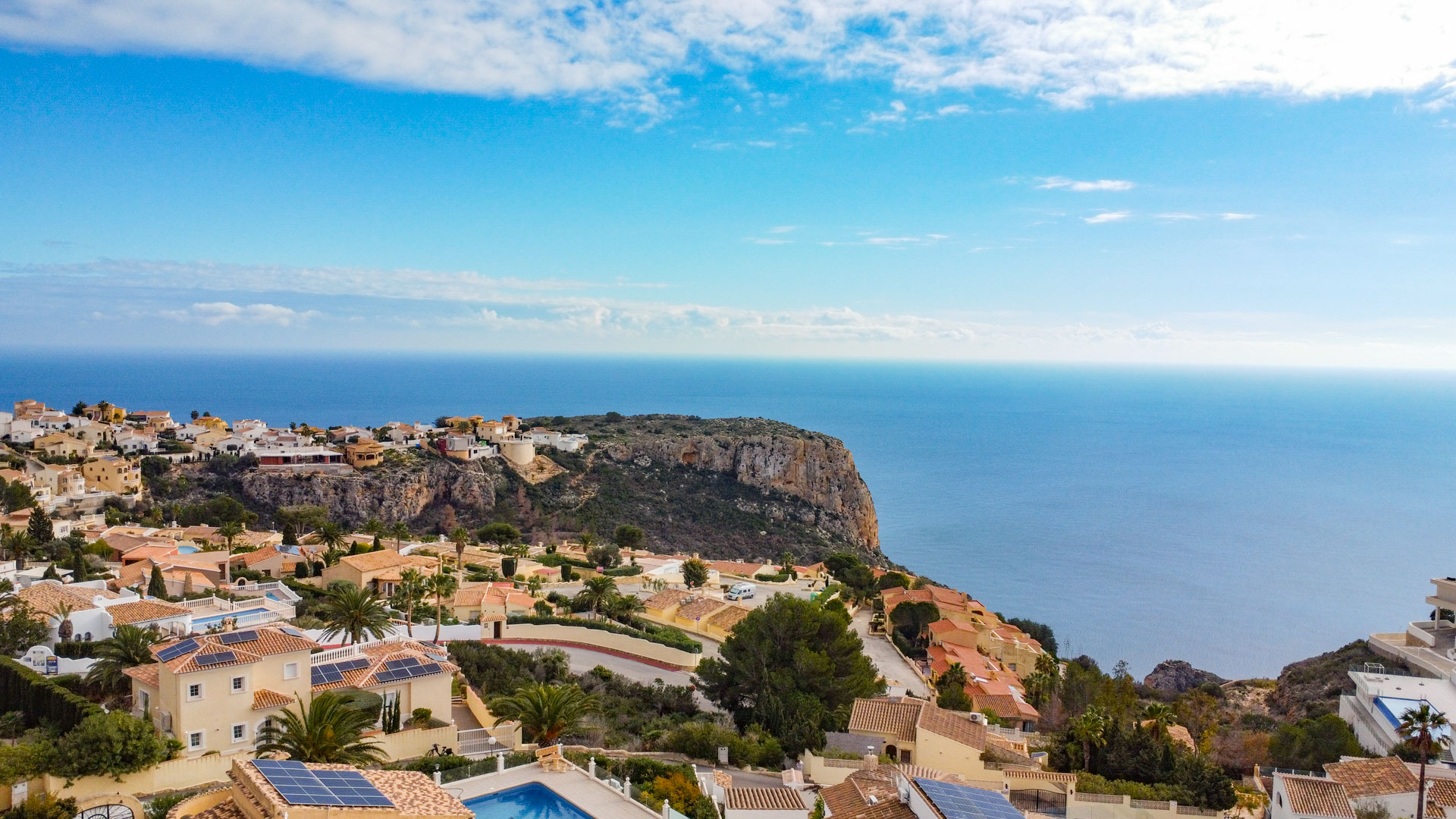 Elevated panoramic view of Cumbre del Sol featuring Mediterranean villas with solar panels, coastal cliffs, and the expansive blue horizon of the sea.